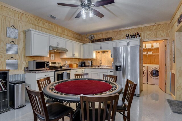 kitchen with wooden walls, ceiling fan, white cabinetry, stainless steel appliances, and washing machine and clothes dryer