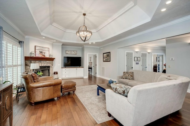 living room with crown molding, a stone fireplace, a notable chandelier, a raised ceiling, and wood-type flooring