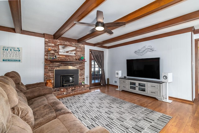 living area featuring baseboards, ceiling fan, beamed ceiling, a wood stove, and wood finished floors