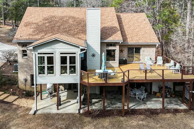 rear view of property featuring a shingled roof, a patio, a chimney, a wooden deck, and brick siding