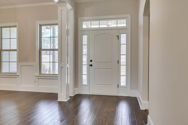 foyer entrance featuring decorative columns, wainscoting, dark wood-style flooring, crown molding, and a decorative wall