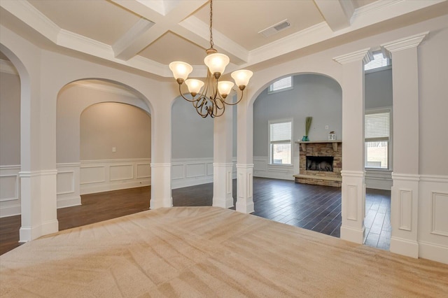 unfurnished dining area with beam ceiling, an inviting chandelier, a stone fireplace, coffered ceiling, and ornate columns