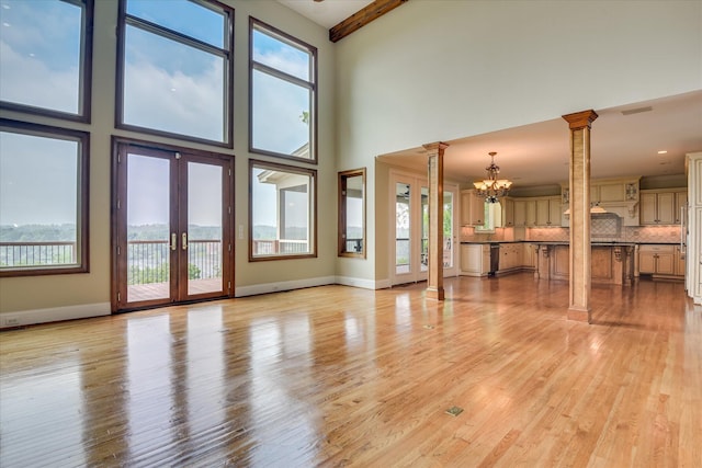 unfurnished living room featuring decorative columns, a chandelier, french doors, light wood-style flooring, and a towering ceiling