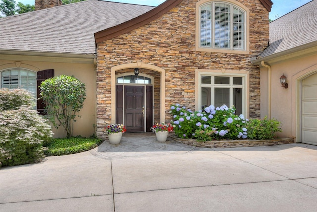 doorway to property with stone siding, roof with shingles, a chimney, stucco siding, and driveway