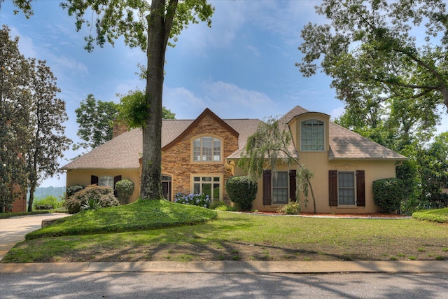traditional home with stucco siding, stone siding, a chimney, a front yard, and a shingled roof