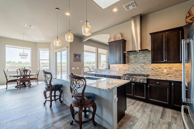 kitchen featuring an island with sink, tasteful backsplash, a breakfast bar area, wall chimney exhaust hood, and light stone counters