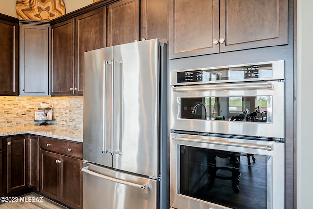 kitchen featuring dark brown cabinetry, light stone counters, stainless steel appliances, light wood-type flooring, and backsplash