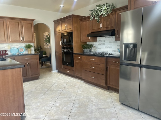 kitchen with dark countertops, under cabinet range hood, light tile patterned flooring, arched walkways, and black appliances