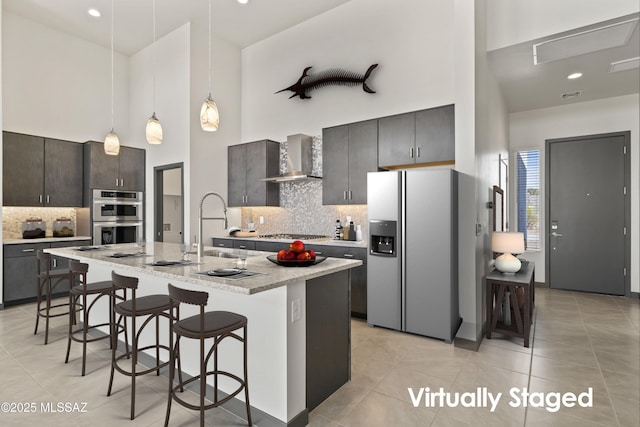 kitchen featuring stainless steel appliances, a breakfast bar, a sink, a towering ceiling, and wall chimney range hood