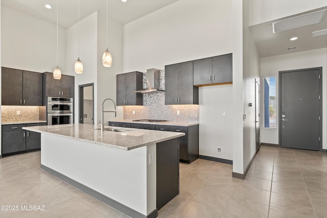 kitchen featuring wall chimney exhaust hood, appliances with stainless steel finishes, a kitchen island with sink, a high ceiling, and a sink