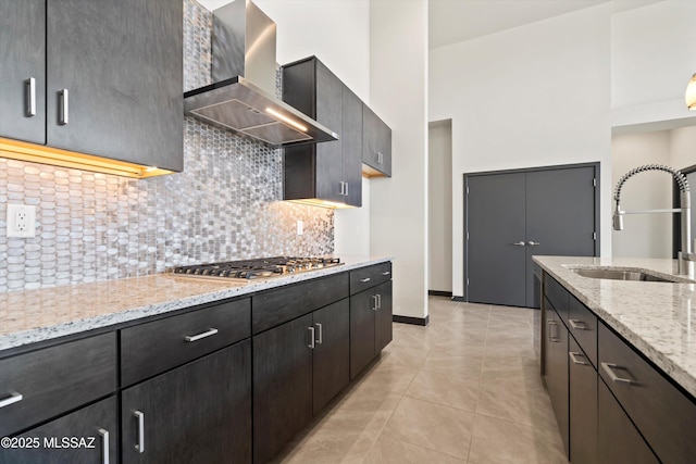 kitchen with stainless steel gas cooktop, a sink, wall chimney range hood, decorative backsplash, and light stone countertops