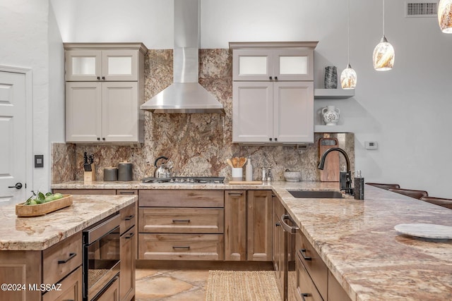 kitchen with wall chimney range hood, sink, light stone countertops, decorative light fixtures, and stainless steel appliances