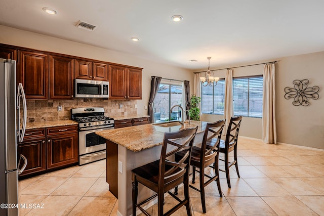 kitchen with light stone counters, an island with sink, a chandelier, decorative light fixtures, and appliances with stainless steel finishes