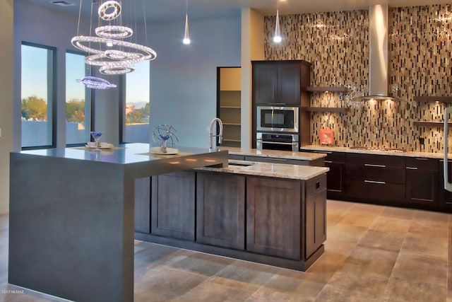 kitchen featuring hanging light fixtures, tasteful backsplash, dark brown cabinetry, a kitchen island with sink, and wall chimney exhaust hood