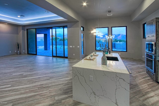 kitchen featuring oven, dark wood-type flooring, a kitchen island with sink, light stone counters, and pendant lighting