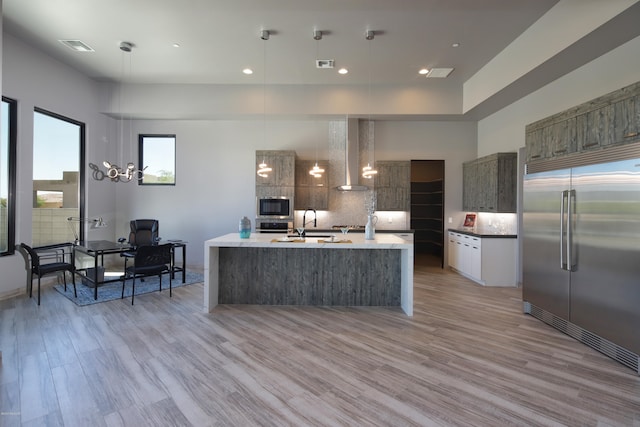 kitchen with appliances with stainless steel finishes, pendant lighting, light wood-type flooring, a center island with sink, and wall chimney exhaust hood