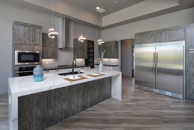 kitchen with wall chimney range hood, an island with sink, stainless steel appliances, backsplash, and light hardwood / wood-style floors