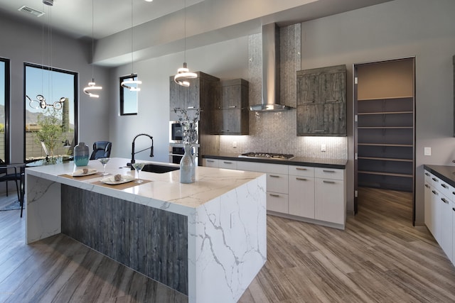 kitchen with a kitchen island with sink, sink, wood-type flooring, wall chimney exhaust hood, and white cabinetry