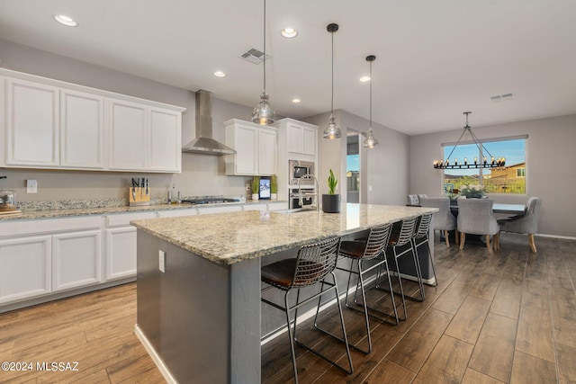 kitchen with light hardwood / wood-style floors, a kitchen island with sink, wall chimney exhaust hood, white cabinetry, and pendant lighting