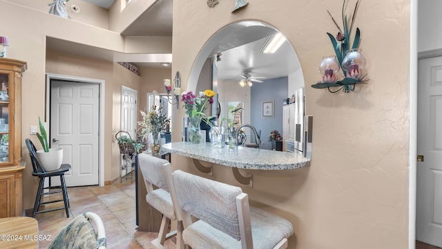 kitchen with light stone counters, ceiling fan, light tile floors, sink, and a breakfast bar area