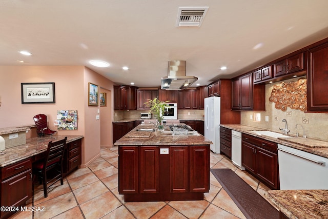 kitchen featuring light stone countertops, island range hood, backsplash, white appliances, and a kitchen island with sink