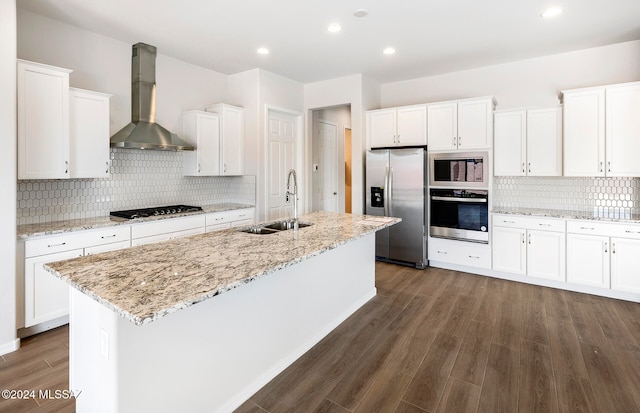 kitchen featuring stainless steel appliances, white cabinets, wall chimney exhaust hood, sink, and a center island with sink