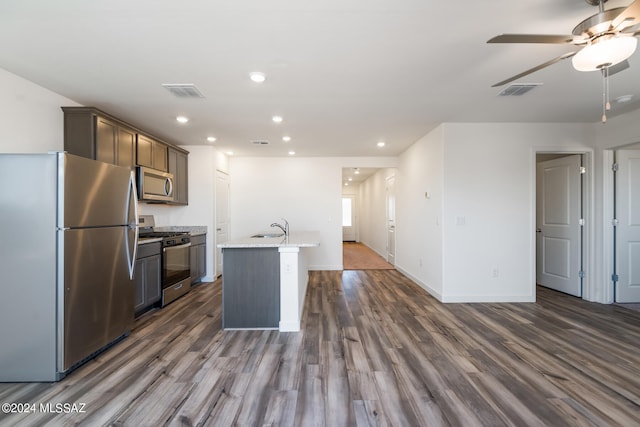 kitchen featuring dark hardwood / wood-style floors, light stone counters, ceiling fan, appliances with stainless steel finishes, and a center island with sink