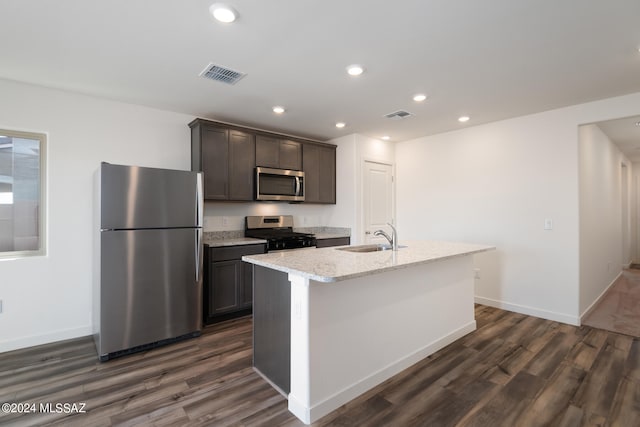 kitchen featuring dark wood-type flooring, appliances with stainless steel finishes, an island with sink, sink, and light stone counters