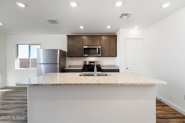 kitchen featuring dark hardwood / wood-style floors, appliances with stainless steel finishes, dark brown cabinetry, light stone countertops, and a center island with sink