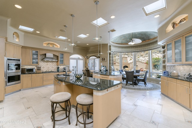 kitchen with wall chimney exhaust hood, hanging light fixtures, a kitchen island with sink, and a skylight