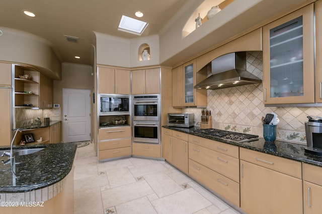 kitchen with appliances with stainless steel finishes, backsplash, a skylight, light brown cabinetry, and wall chimney range hood