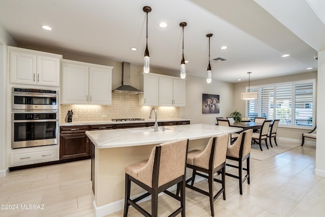 kitchen featuring sink, wall chimney exhaust hood, hanging light fixtures, a kitchen island with sink, and white cabinets