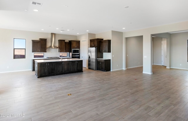 kitchen featuring a kitchen island with sink, stainless steel appliances, wall chimney exhaust hood, light hardwood / wood-style floors, and backsplash