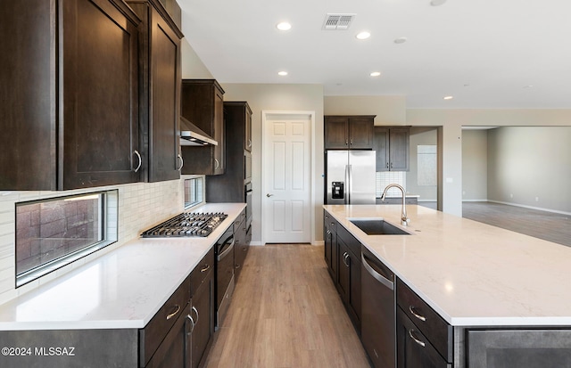 kitchen with light wood-type flooring, sink, decorative backsplash, an island with sink, and appliances with stainless steel finishes