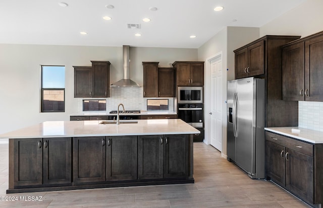 kitchen with wall chimney exhaust hood, a center island with sink, appliances with stainless steel finishes, and light wood-type flooring