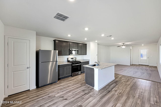 kitchen with stainless steel appliances, sink, an island with sink, ceiling fan, and light wood-type flooring