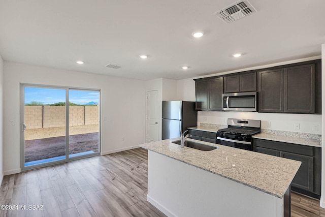 kitchen with light hardwood / wood-style floors, sink, appliances with stainless steel finishes, light stone countertops, and an island with sink