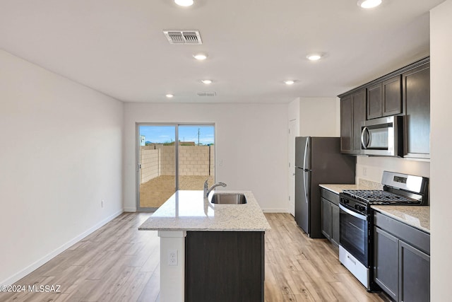 kitchen with stainless steel appliances, sink, light stone counters, an island with sink, and light wood-type flooring