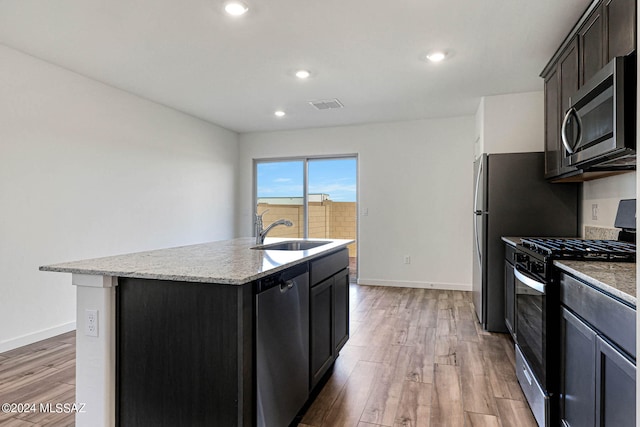 kitchen with light wood-type flooring, appliances with stainless steel finishes, light stone countertops, sink, and a kitchen island with sink