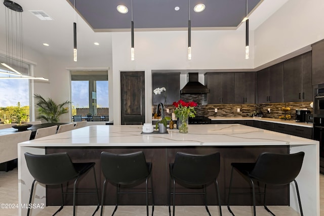 kitchen featuring wall chimney range hood, a spacious island, tasteful backsplash, light stone countertops, and decorative light fixtures