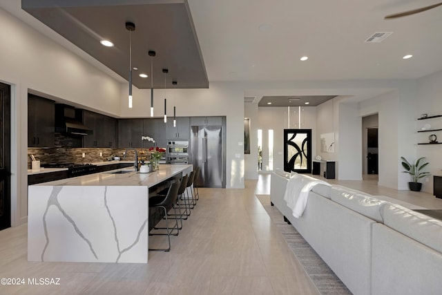 kitchen featuring sink, hanging light fixtures, a large island with sink, decorative backsplash, and wall chimney exhaust hood