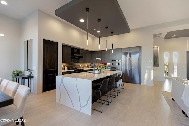 kitchen with appliances with stainless steel finishes, sink, backsplash, a kitchen island with sink, and wall chimney range hood