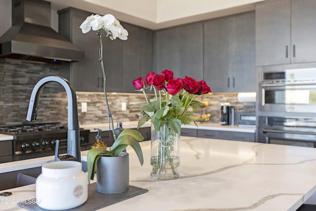 interior space with light stone counters, tasteful backsplash, gray cabinets, stainless steel double oven, and wall chimney range hood