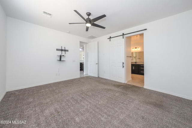 unfurnished bedroom featuring light colored carpet, a barn door, ceiling fan, and ensuite bathroom