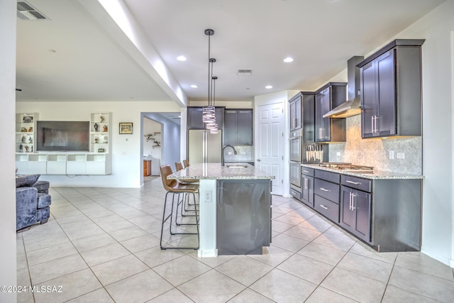 kitchen with pendant lighting, sink, light stone countertops, an island with sink, and wall chimney exhaust hood