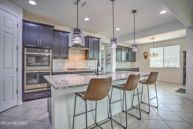 kitchen featuring pendant lighting, appliances with stainless steel finishes, a center island with sink, light stone countertops, and a kitchen bar