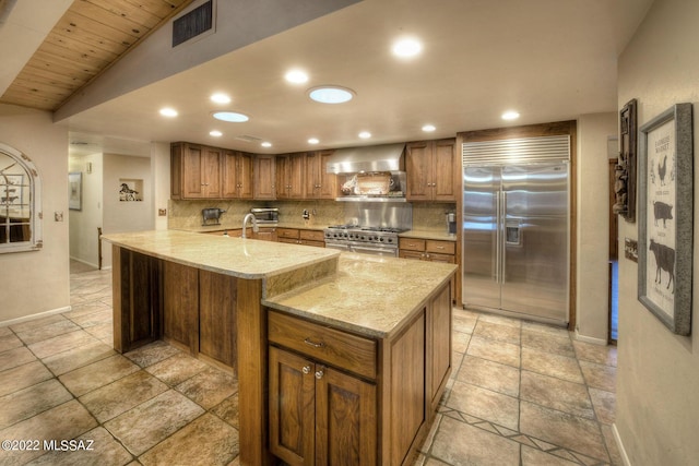 kitchen with high end appliances, wall chimney range hood, decorative backsplash, light stone counters, and kitchen peninsula