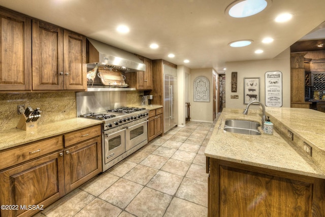 kitchen featuring backsplash, light stone counters, wall chimney exhaust hood, sink, and range with two ovens