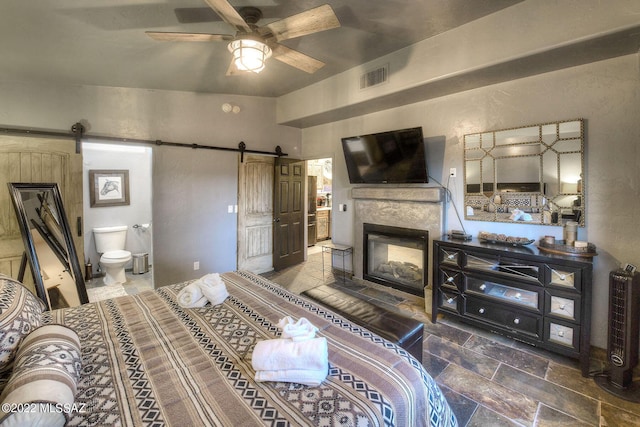 bedroom featuring a barn door, ceiling fan, ensuite bath, and a tiled fireplace