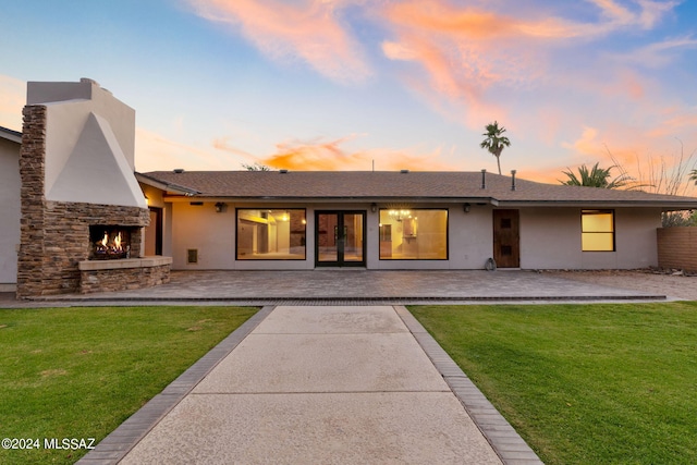 back of property at dusk featuring stucco siding, an outdoor stone fireplace, french doors, a patio area, and a lawn